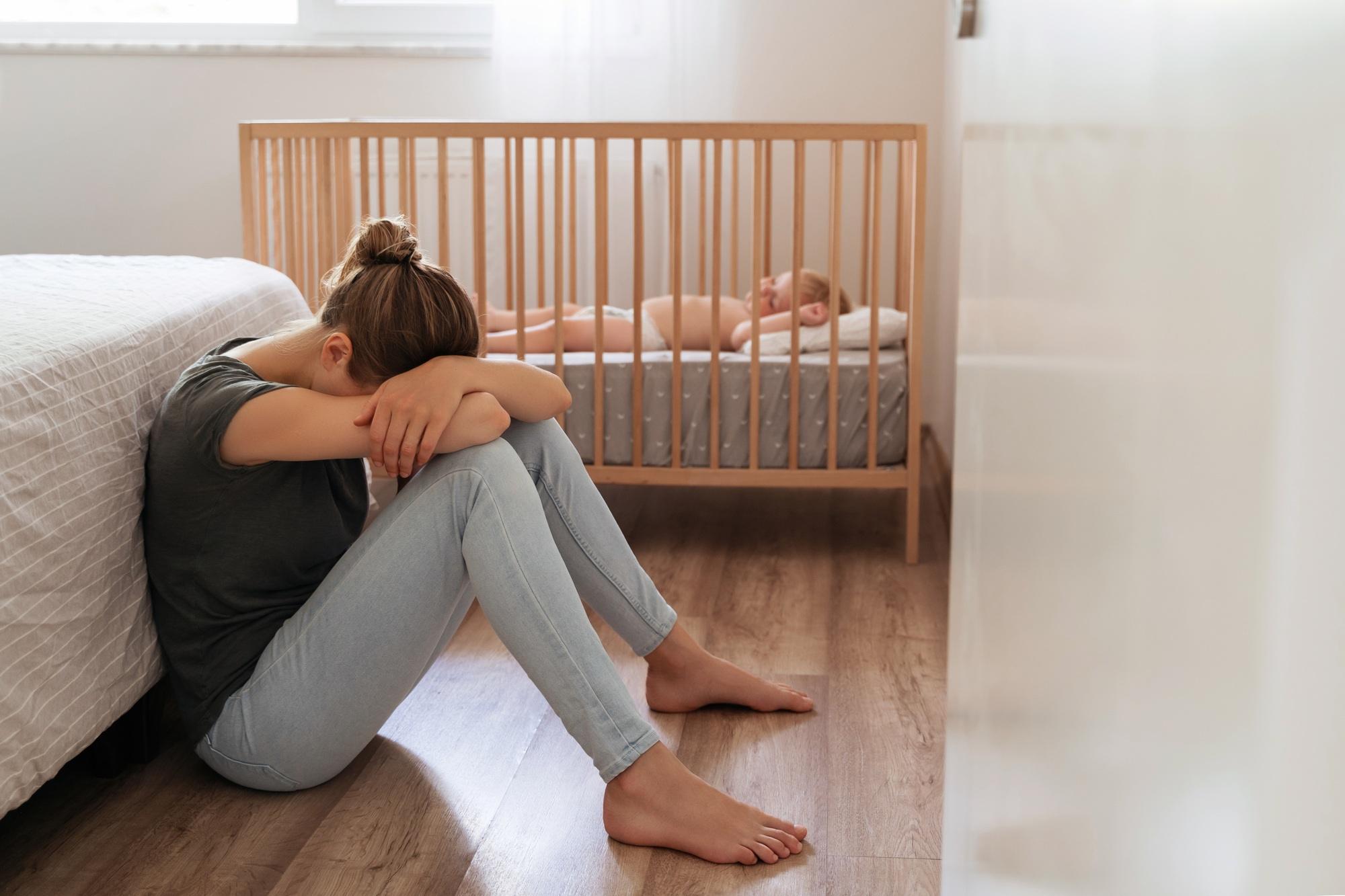 Mother suffering postnatal depression sitting on floor with head on knees next to baby crib