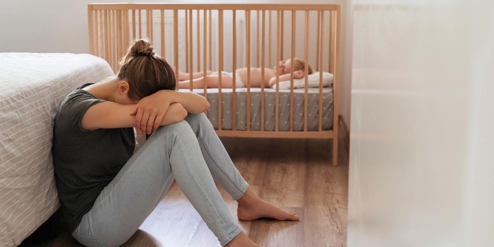 Mother suffering postnatal depression sitting on floor with head on knees next to baby crib