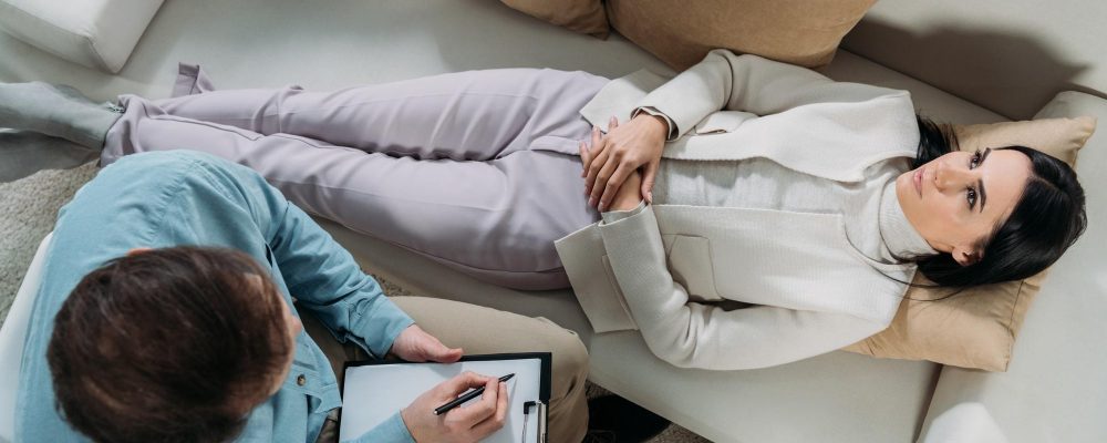 overhead view of psychotherapist writing on clipboard and upset young patient lying on couch